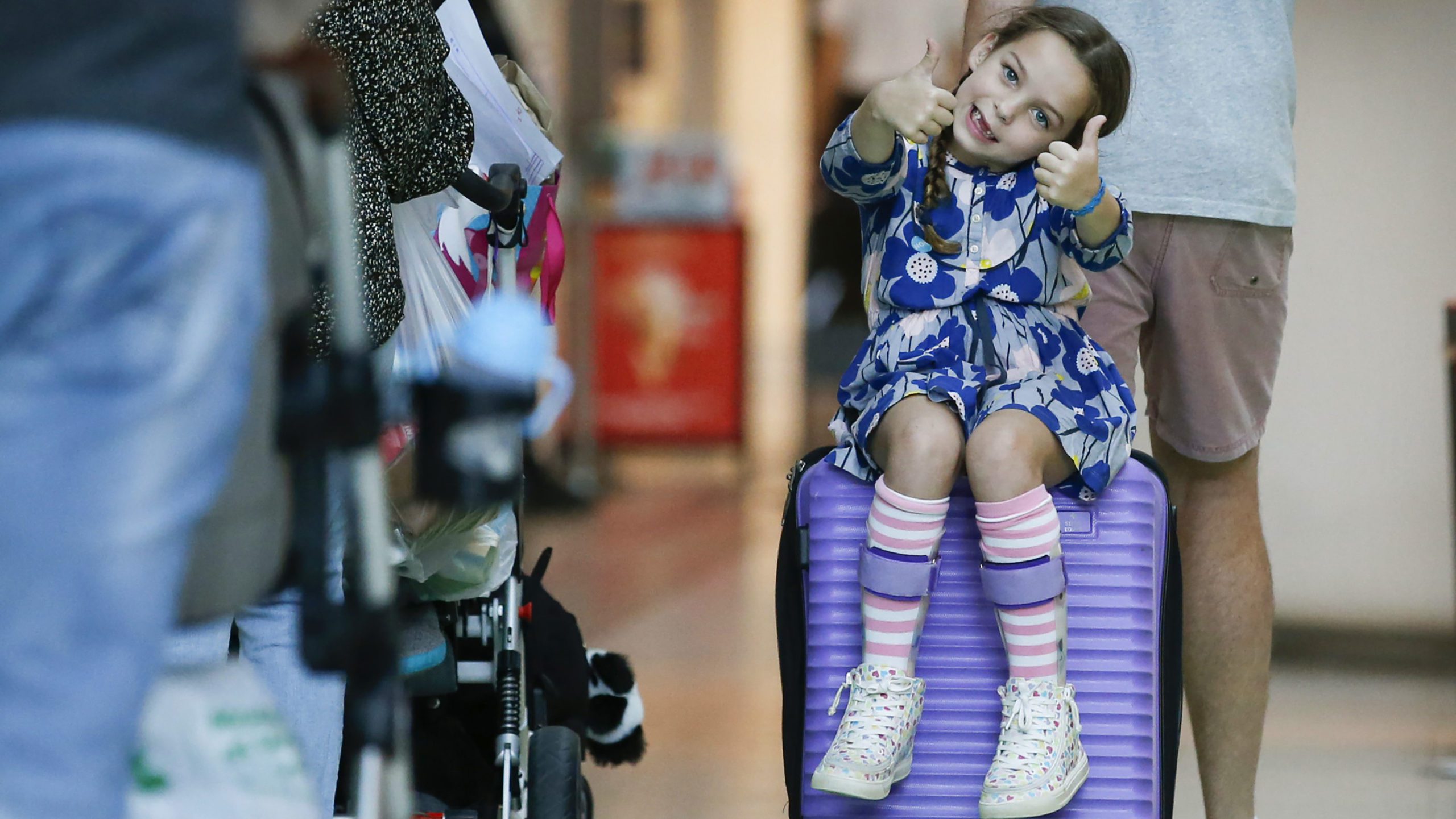 Piper is sitting on her purple suitcase, holding her her thumbs up