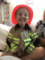 Stella sitting on her hospital bed wearing a costume firefighter jacket and helmet. She has a nasogastric tube in her nose