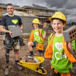 A man with a prosthetic leg holding bricks and his two kids holding bricks standing in front of the charity house which is under construction