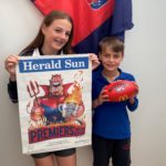 A girl holding a Melbourne Premiership Poster with a demon caricature on it and a boy holding a football. They are standing in front of a Melbourne FC flag