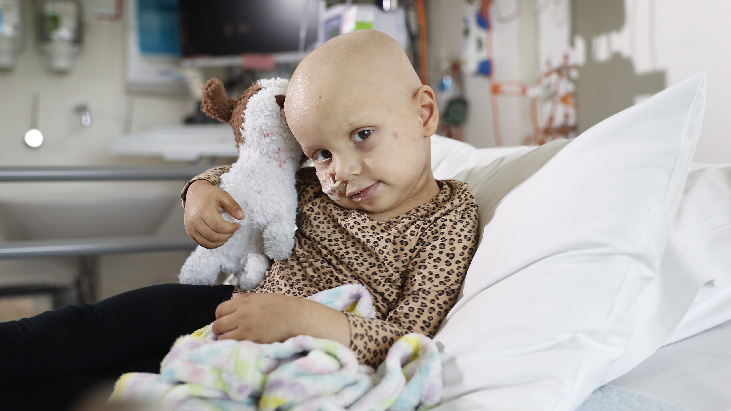 Ayla sitting on her hospital bed holding her stuffed dog toy. Ayla is bald and has a nasal gastic tube.