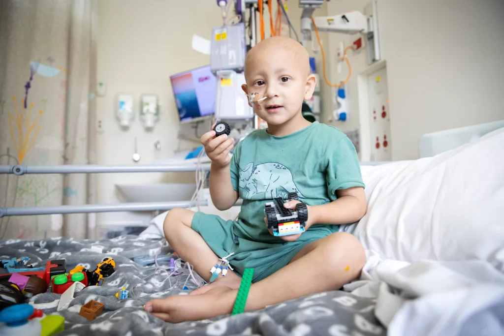 Ibrahim sits on a hospital bed at The Royal Childrens Hospital Melbourne in a light blue shirt playing with toy trucks and cars.